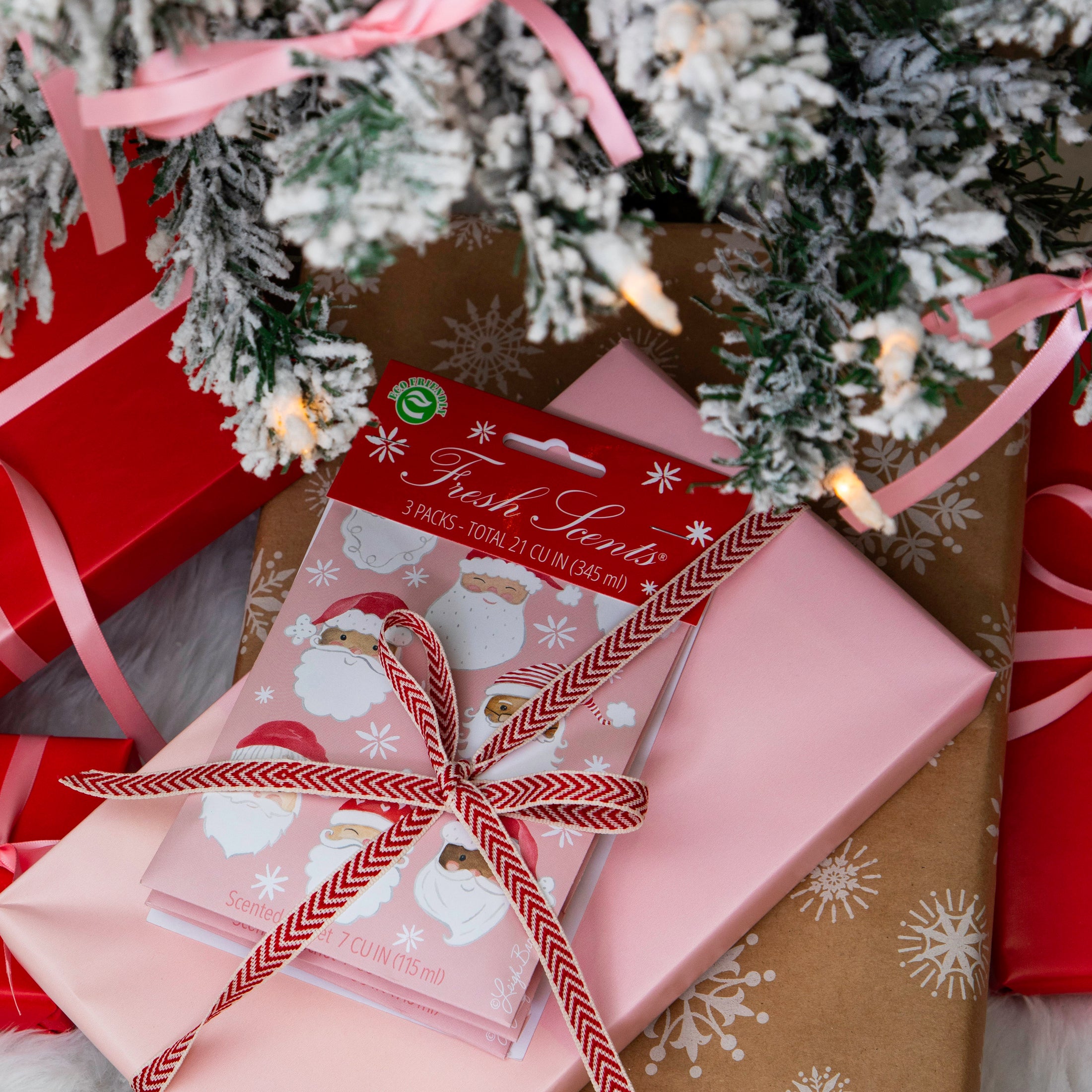 Pack of pink scented sachets with watercolor santas on it displayed under a frosted Christmas tree wrapped in a bow with other gifts.