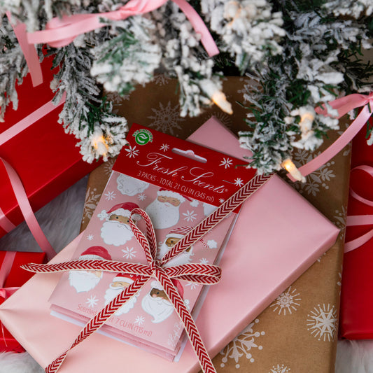 Pack of pink scented sachets with watercolor santas on it displayed under a frosted Christmas tree wrapped in a bow with other gifts.