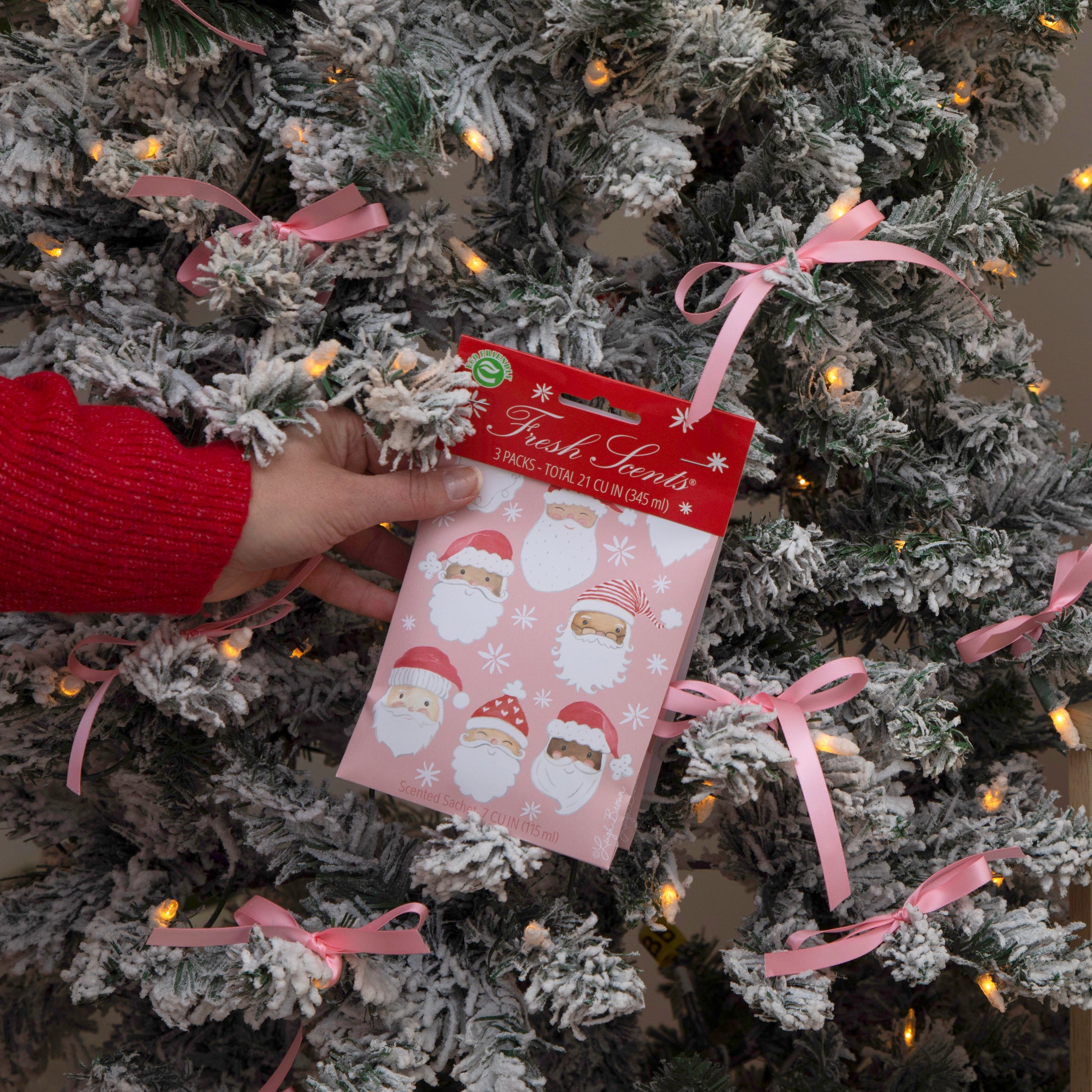 Pack of pink scented sachets with watercolor santas on it being held by a hand in front of a frosted Christmas Tree