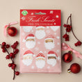 Load image into Gallery viewer, Pack of pink scented sachets with watercolor santas on it displayed on a tan background with red ornaments and berries.