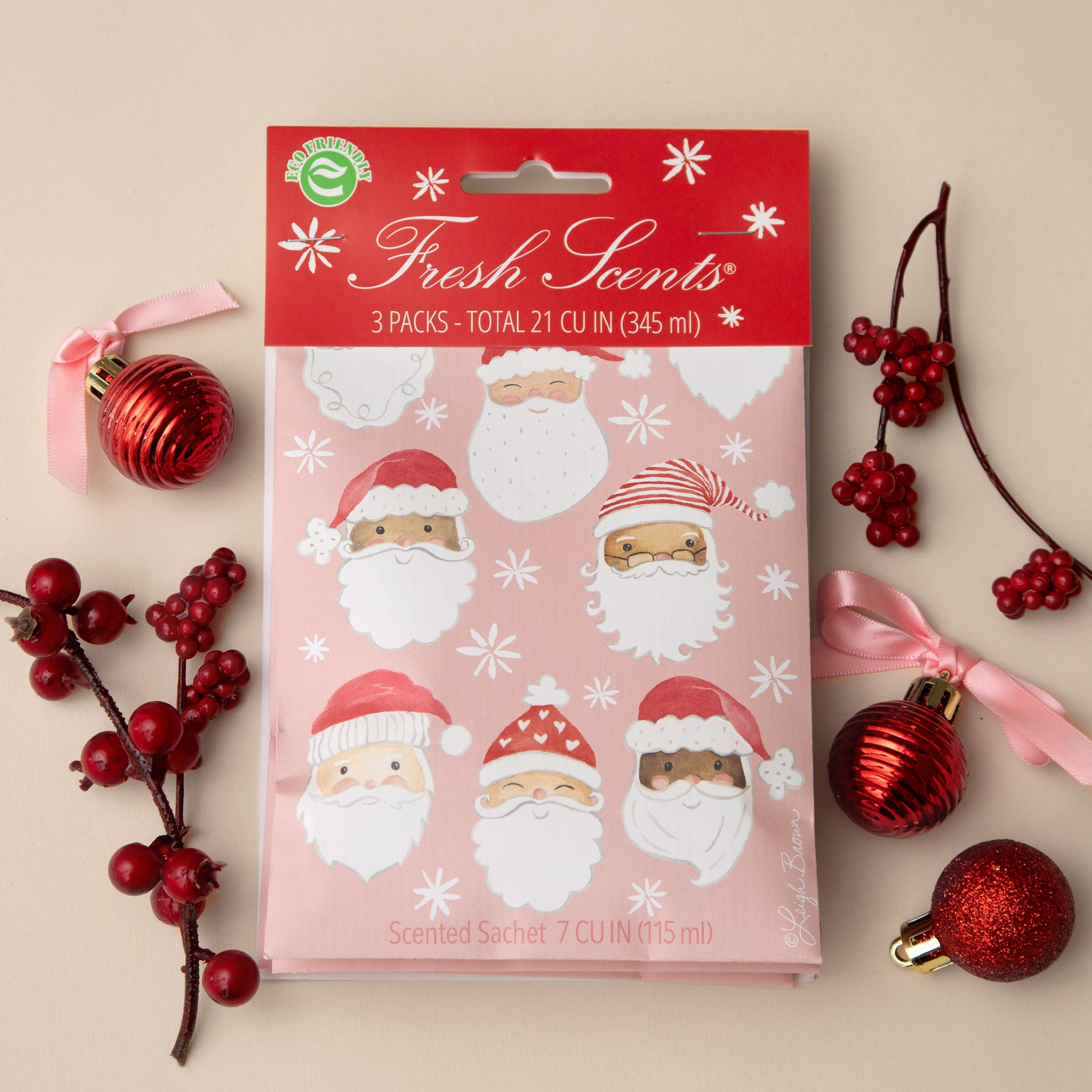 Pack of pink scented sachets with watercolor santas on it displayed on a tan background with red ornaments and berries.