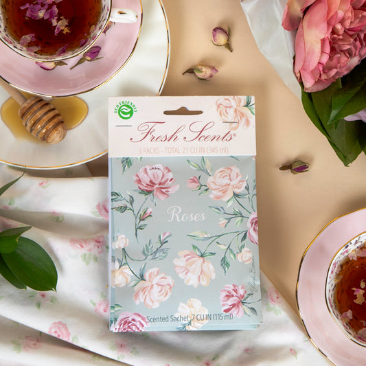 A pack of scented sachets with a floral design, labeled 'Roses', displayed on a table with a cup of tea and roses in the background.