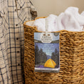 Load image into Gallery viewer, A woven laundry basket filled with white towels features a Fresh Scents Into the Forest - Sachet 3pk for a long-lasting fragrance, while a plaid shirt hangs on the left side.