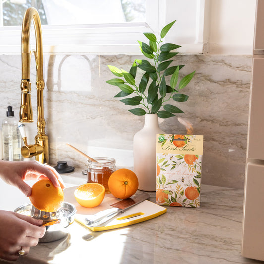 A scented sachet pack with water color oranges on it being displayed on a kitchen countertop while a hand juices oranges.