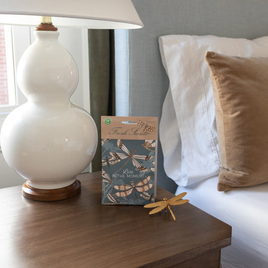 A pack of three scented sachets with a dragonfly design and the phrase 'Live in the Moment' on the packaging, displayed on a nightstand with a dragonfly paperweight and a lamp.