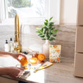 Load image into Gallery viewer, A scented sachet pack with water color oranges on it being displayed on a kitchen countertop while a hand juices oranges.