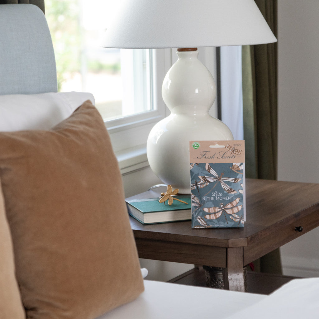 A pack of three scented sachets with a dragonfly design and the phrase 'Live in the Moment' on the packaging, displayed on a nightstand with a book and a lamp