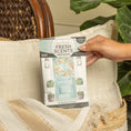 Load image into Gallery viewer, A hand holds the Fresh Scents Spring Door - Sachet 3pk, a long-lasting flameless freshener featuring a pastel blue door and floral wreath design, over a cream pillow and woven basket, beside a green plant.