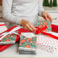 Load image into Gallery viewer, Wearing a white sweater, a person adds Peace on Earth - Sachet 3pk by Fresh Scents to red and white wrapped gifts on a table, with green decor in the background.