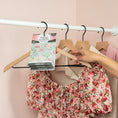 Load image into Gallery viewer, A hand hangs a floral dress on a wooden hanger in a closet, where several other garments and hangers are seen along with Fresh Scents Roses - Sachet 3pk for long-lasting freshness hanging on the closet rod.