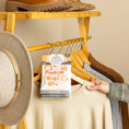Load image into Gallery viewer, A hand hangs a Fresh Scents You're the Pumpkin - Sachet 3pk on a wooden rack with sweaters, a tan hat, and brown shoes—a charming flameless air freshener for long-lasting fragrance.
