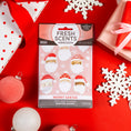 Load image into Gallery viewer, A festive flat lay displays the Fresh Scents Merry Santas - Sachet 3pk, featuring cartoon Santa faces, ornaments, and gifts—enjoy this flameless, long-lasting air freshener on a vibrant red background.