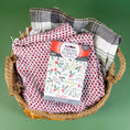 Load image into Gallery viewer, A woven basket holds two folded dish towels—one red and white, one green and white—with a Fresh Scents Be Merry - Sachet 3pk on top, set against a green background.