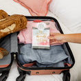 Load image into Gallery viewer, A hand packs a Fresh Scents She Believed - Sachet 3pk into a suitcase with clothes like jeans, a pink striped shirt, and cowboy boots—perfect as a long-lasting air freshener for travel.