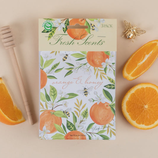 A pack of three scented sachets with a orange and honey design, accompanied by a wooden honey dipper and slices of orange.