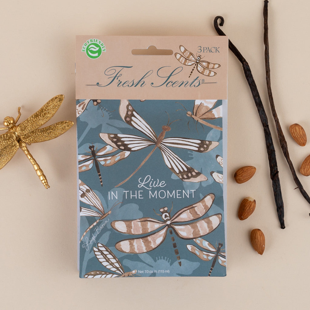 A pack of three scented sachets with a dragonfly design and the phrase 'Live in the Moment' on the packaging, accompanied by vanilla beans and almonds on a beige background.