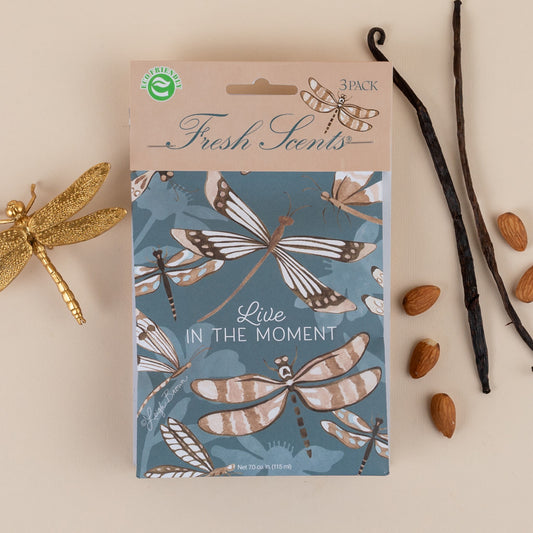A pack of three scented sachets with a dragonfly design and the phrase 'Live in the Moment' on the packaging, accompanied by vanilla beans and almonds on a beige background.