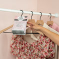 Load image into Gallery viewer, A hand hangs a Fresh Scents Faith Hope Love - Sachet 3pk, a flameless floral air freshener, on a wooden hanger between floral and light-colored clothes in a closet with pink walls and a white rod.