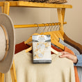 Load image into Gallery viewer, A person hangs a beige sweater on a wooden rack with several clothes and a beige hat, while a Fresh Scents Fall & Family - Sachet 3pk flameless air freshener sits in front.