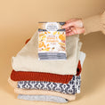 Load image into Gallery viewer, A hand holds a Fresh Scents Autumn Leaves - Sachet 3pk above a stack of folded sweaters in cozy fall colors and patterns, set against a beige background.