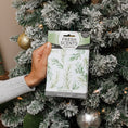Load image into Gallery viewer, A hand holds a Fresh Scents Balsam Fir - Sachet 3pk in front of a decorated, snow-frosted Christmas tree with gold and silver ornaments, adding long-lasting freshness and flameless fragrance to your holiday home.