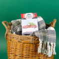 Load image into Gallery viewer, A woven basket holds a plaid fringed blanket and a Fresh Scents Merry Christmas - Sachet 3pk featuring a red truck and tree design, providing long-lasting flameless fragrance, set against a green background.