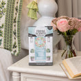 Load image into Gallery viewer, A Fresh Scents Spring Door - Sachet 3pk sits on a white bedside table beside pink roses, baby's breath, an open book, and a lamp. This flameless fragrance freshens any space. In the background is a green embroidered pillow.
