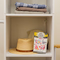 Load image into Gallery viewer, A closet shelf holds folded clothes, while the lower shelf displays a straw hat and Fresh Scents Happy Chicken - Sachet 3pk flameless air fresheners for long-lasting fragrance.