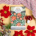 Load image into Gallery viewer, The Peace on Earth - Sachet 3pk by Fresh Scents rests on a tan surface surrounded by red poinsettias, pine branches, a cheerful red mushroom decoration, and a cozy knit red and white blanket. Featuring festive floral designs and labeled with “Peace on Earth,” these scented sachets make the perfect flameless fragrance and long-lasting freshener, beautifully infusing your space with holiday charm.