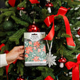 Load image into Gallery viewer, A hand holds a Fresh Scents Peace on Earth - Sachet 3pk package, featuring flameless fragrance sachets, in front of a Christmas tree decorated with red bows, ornaments, and a white snowflake ornament.