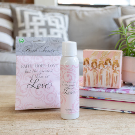 A product image featuring a 3-pack of sachets and a room spray bottle, both with the 'Faith Hope Love' fragrance. The sachets are pink with floral patterns and the spray bottle is white with a pink label, displayed on a coffee table with an artwork and books.