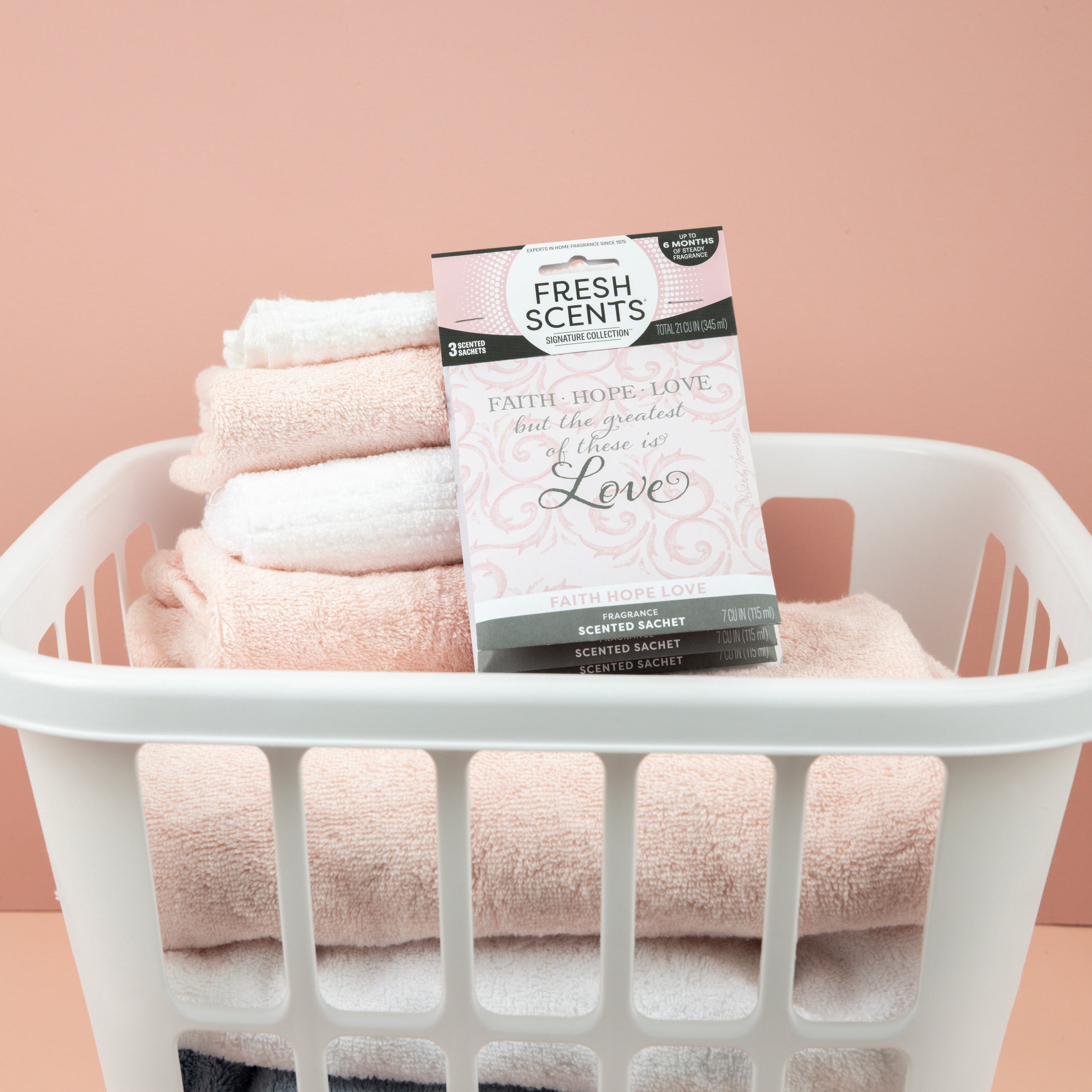 A white laundry basket with folded pastel towels sits before a pink background. On top is a Fresh Scents Faith Hope Love - Sachet 3pk for long-lasting fragrance.