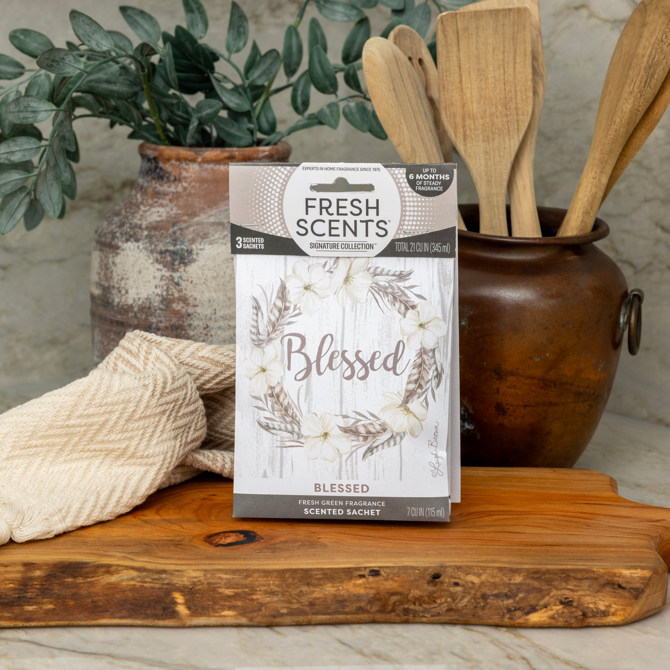 A box labeled Fresh Scents Blessed Wreath 3pk sits on a wooden board, offering flameless fragrance, with a tan towel, wooden utensils in a ceramic pot, and green foliage in the background on a kitchen counter.
