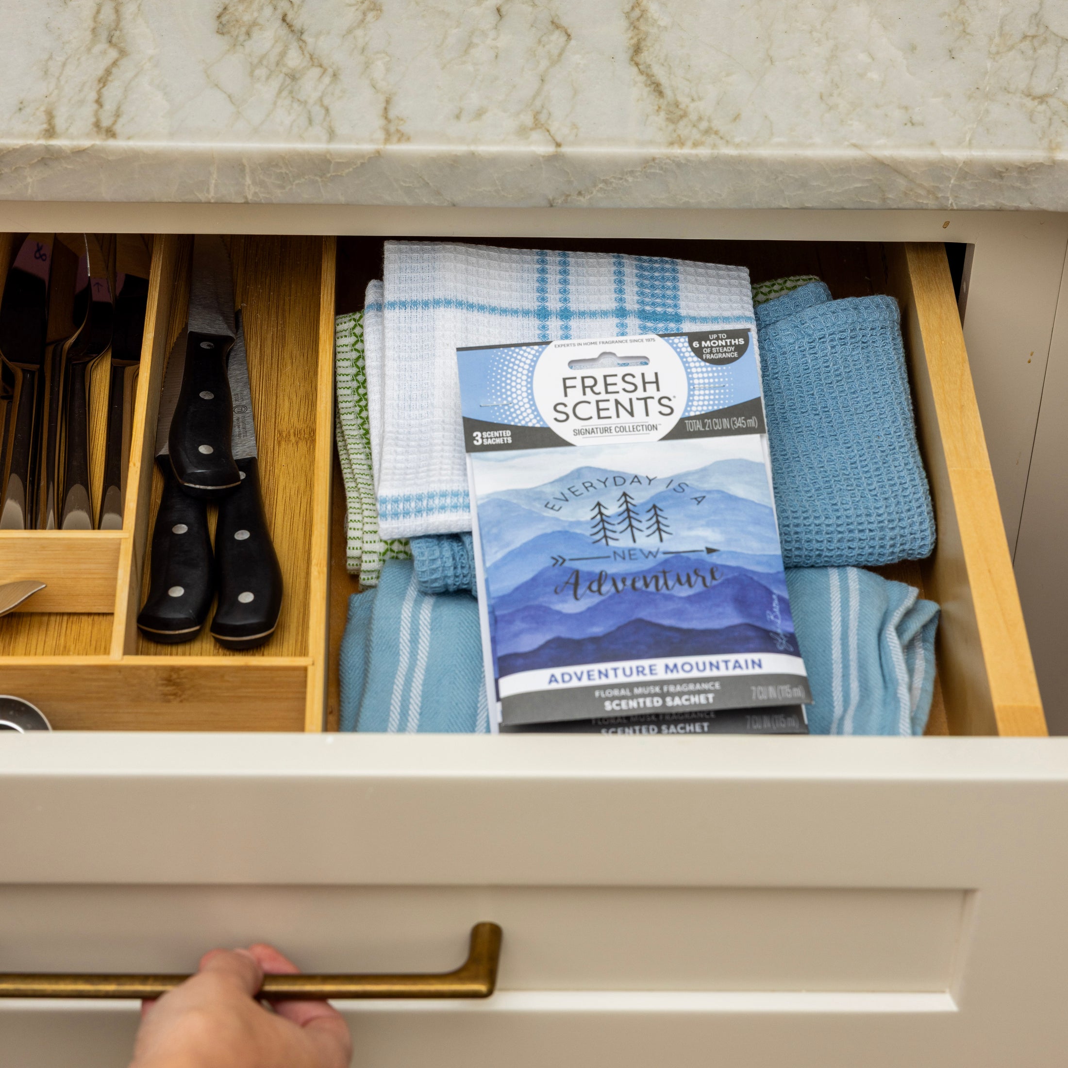 An open kitchen drawer holds folded blue and white dish towels, utensils in a wooden organizer, and a Fresh Scents Adventure Mountain Sachet 3pk for long-lasting freshness. A hand pulls the drawer open beneath a marble countertop.