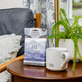 Load image into Gallery viewer, A small table holds a white mug, a potted fern, and a blue bag of Fresh Scents Adventure Mountain - Sachet 3pk for long-lasting freshness. In the background are a gray pillow, wooden chair, and floral curtain by the window.
