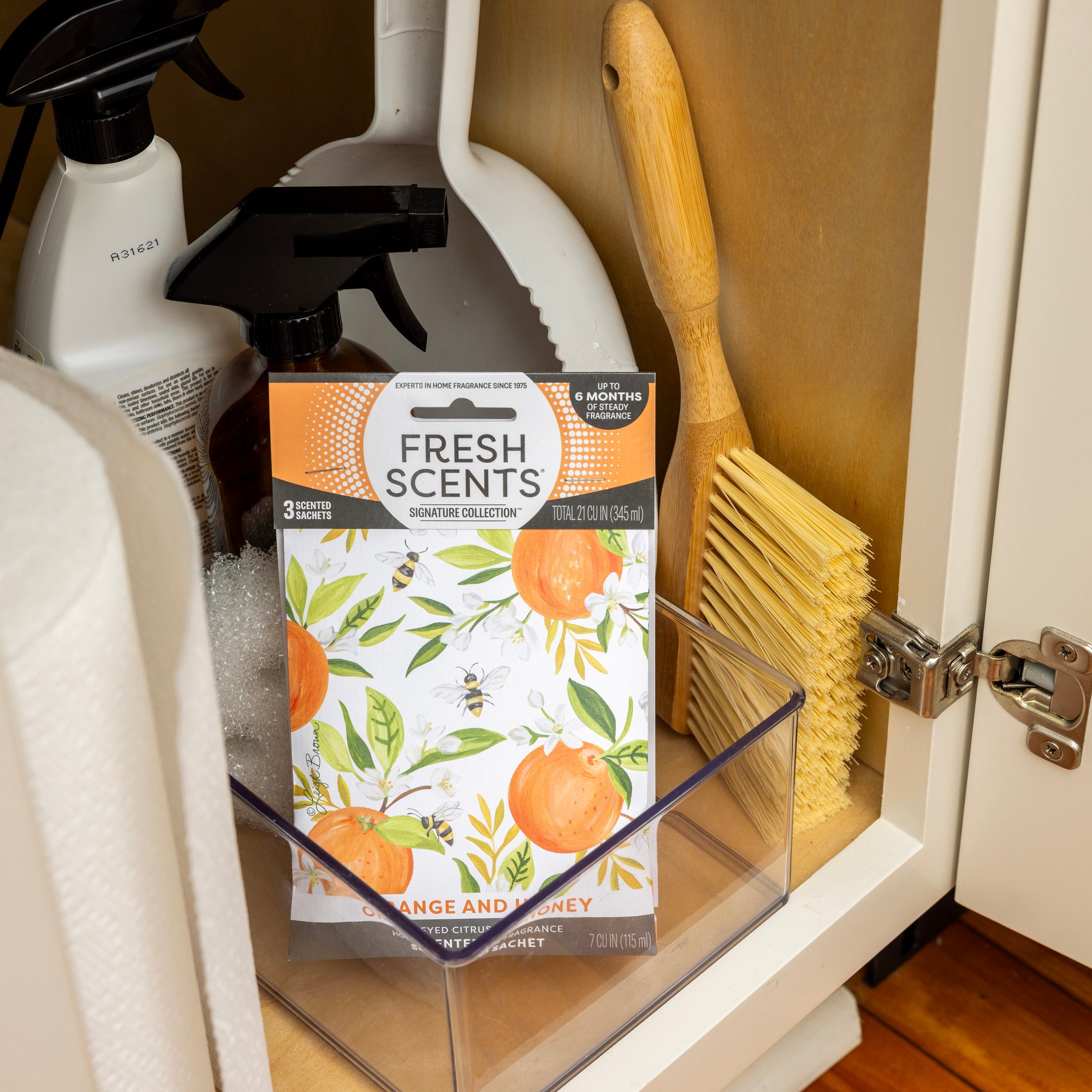 A Fresh Scents Orange & Honey - Sachet 3pk rests in a clear bin inside a cabinet with cleaning sprays and a wooden brush, next to a partially open white door for easy flameless fragrance.