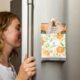 Load image into Gallery viewer, A woman smells the Fresh Scents Orange & Honey - Sachet 3pk, featuring an orange fruit floral design and ribbon for hanging on the fridge. This flameless freshener offers a long-lasting scent for your space.
