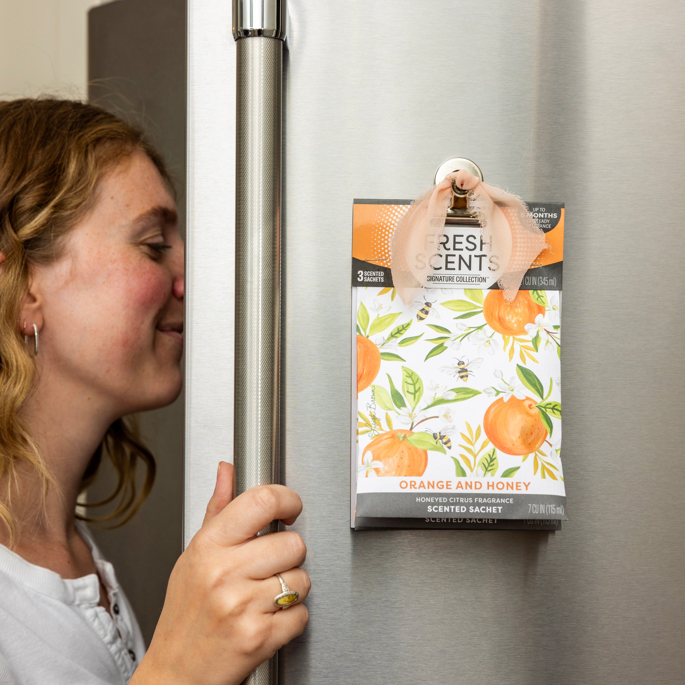 A woman smells the Fresh Scents Orange & Honey - Sachet 3pk, featuring an orange fruit floral design and ribbon for hanging on the fridge. This flameless freshener offers a long-lasting scent for your space.