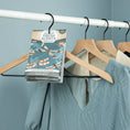 Load image into Gallery viewer, Several wooden hangers with clothes hang on a white closet rod. A Fresh Scents Live in the Moment - Sachet 3pk hangs on one hanger, suggesting lasting freshness for your wardrobe. The background wall is light blue.
