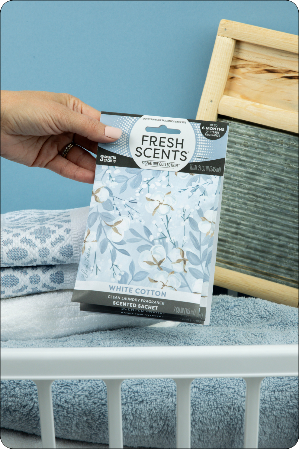 Hand holding a White Cotton 'Fresh Scents' scented sachet package over a laundry basket with blue towels.
