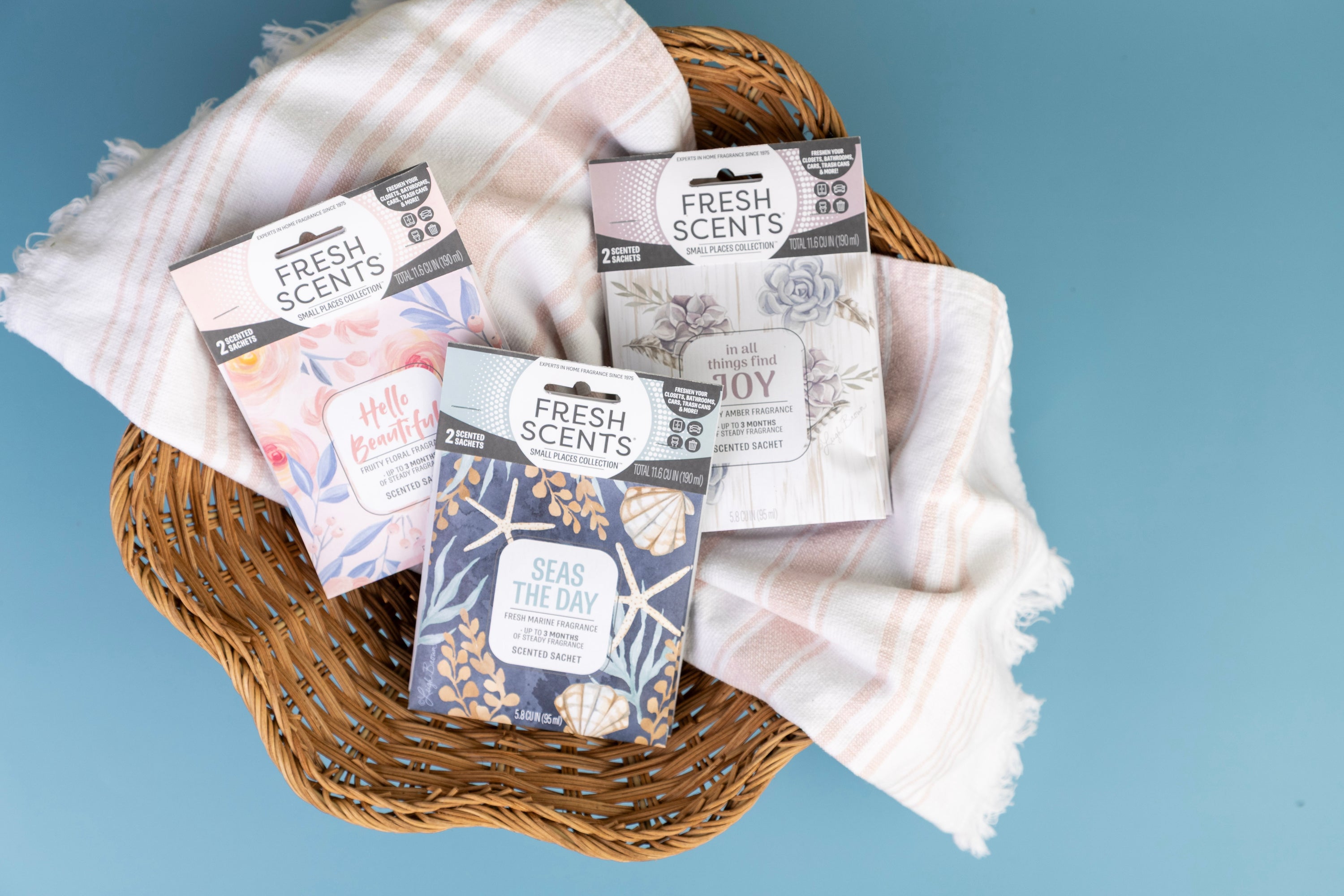 Three Fresh Scents sachet packets in different designs are arranged in a wicker basket on top of a folded, light pink and white striped cloth, set against a blue background.