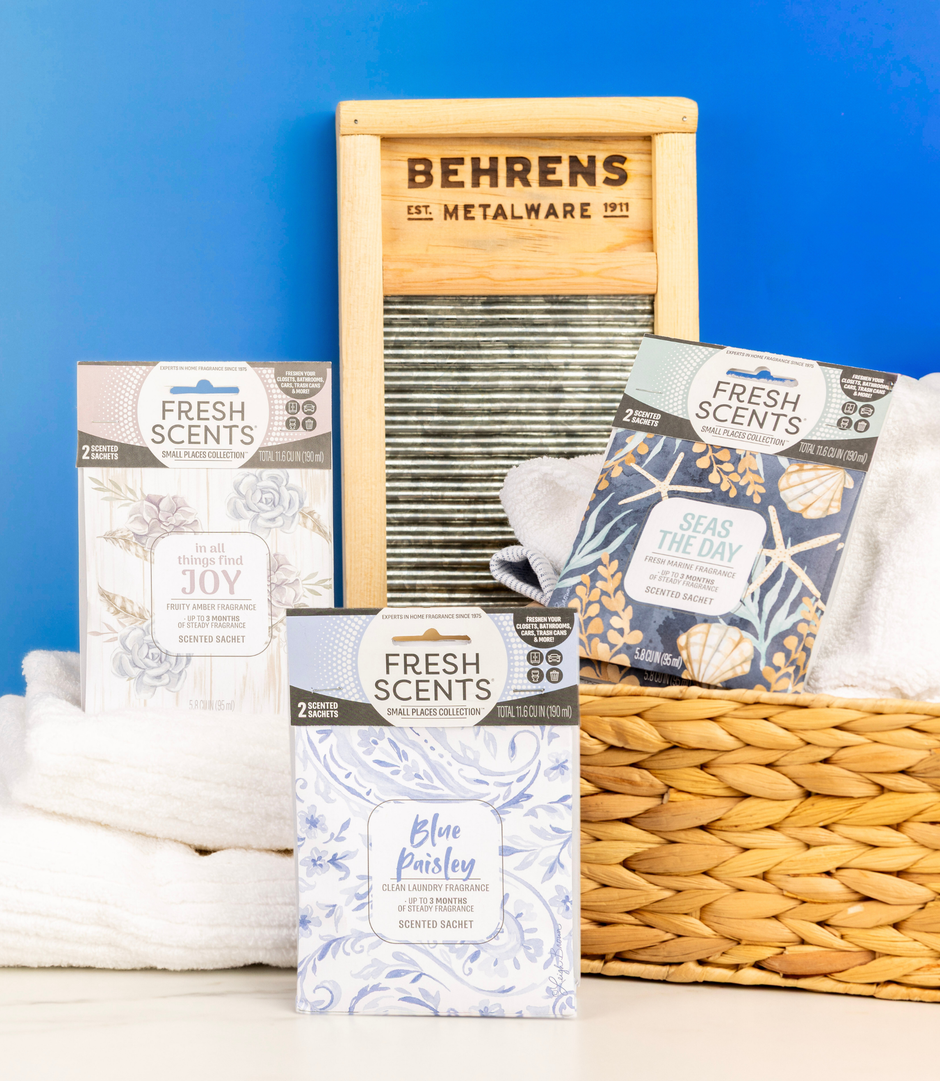 Three Fresh Scents scented sachets are displayed with folded white towels, a wooden washboard, and a woven basket against a blue background. The sachet designs include floral and paisley patterns.