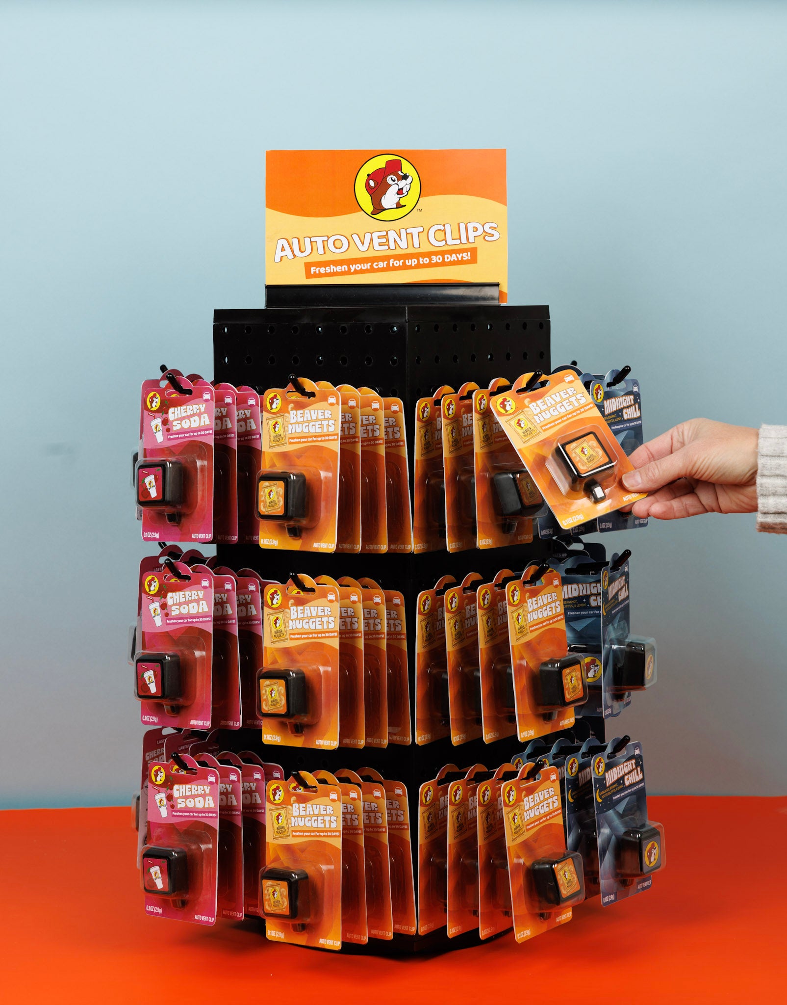 A rotating display stand holds multiple rows of Buc-ee’s auto vent clips in various scents, including Cherry Soda and Beaver Nuggets. A hand reaches to pick up one of the clips.