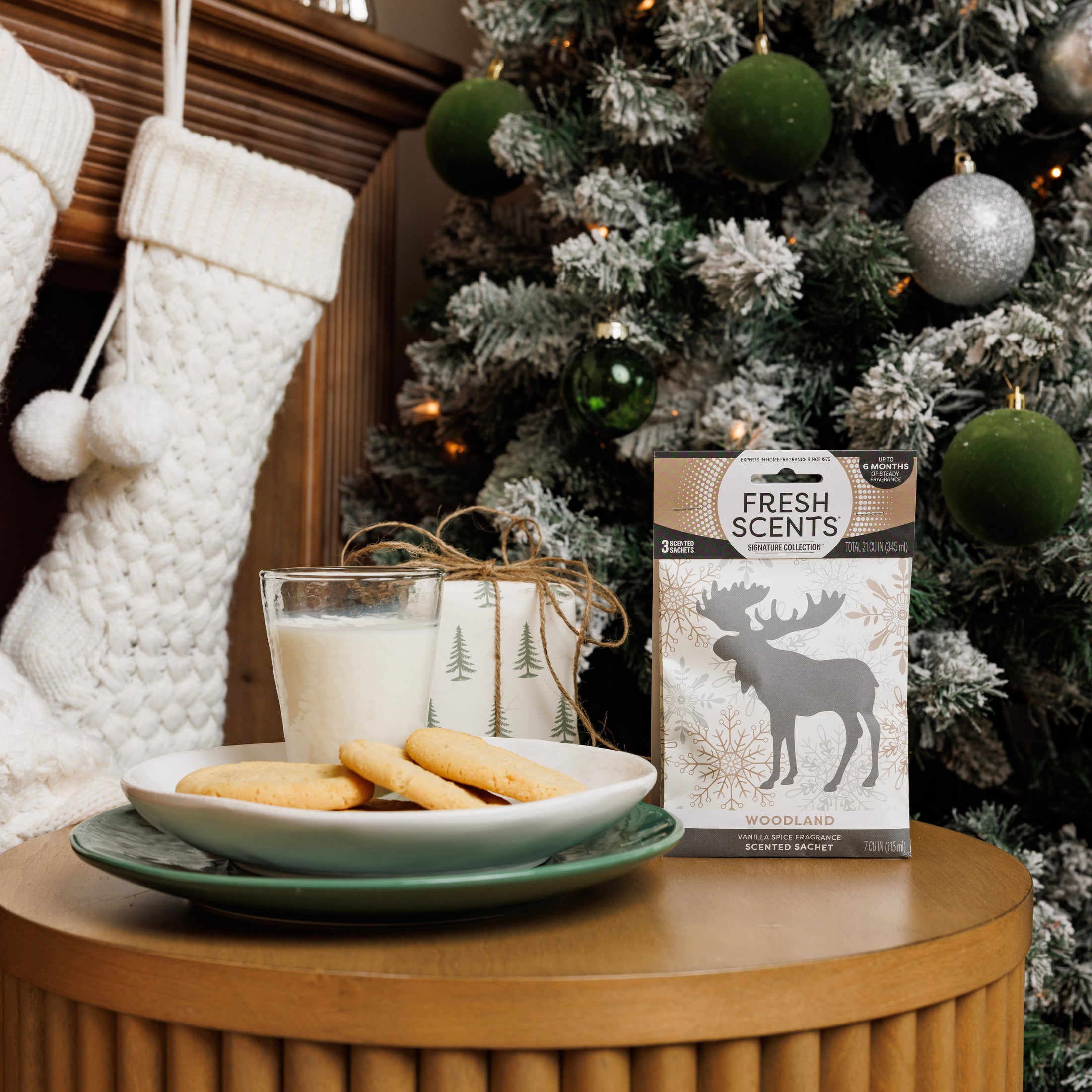 A table with cookies, milk, tree-designed mugs, and a Fresh Scents Woodland Sachet 3pk sits in front of a Christmas tree and white stockings hanging on the fireplace.