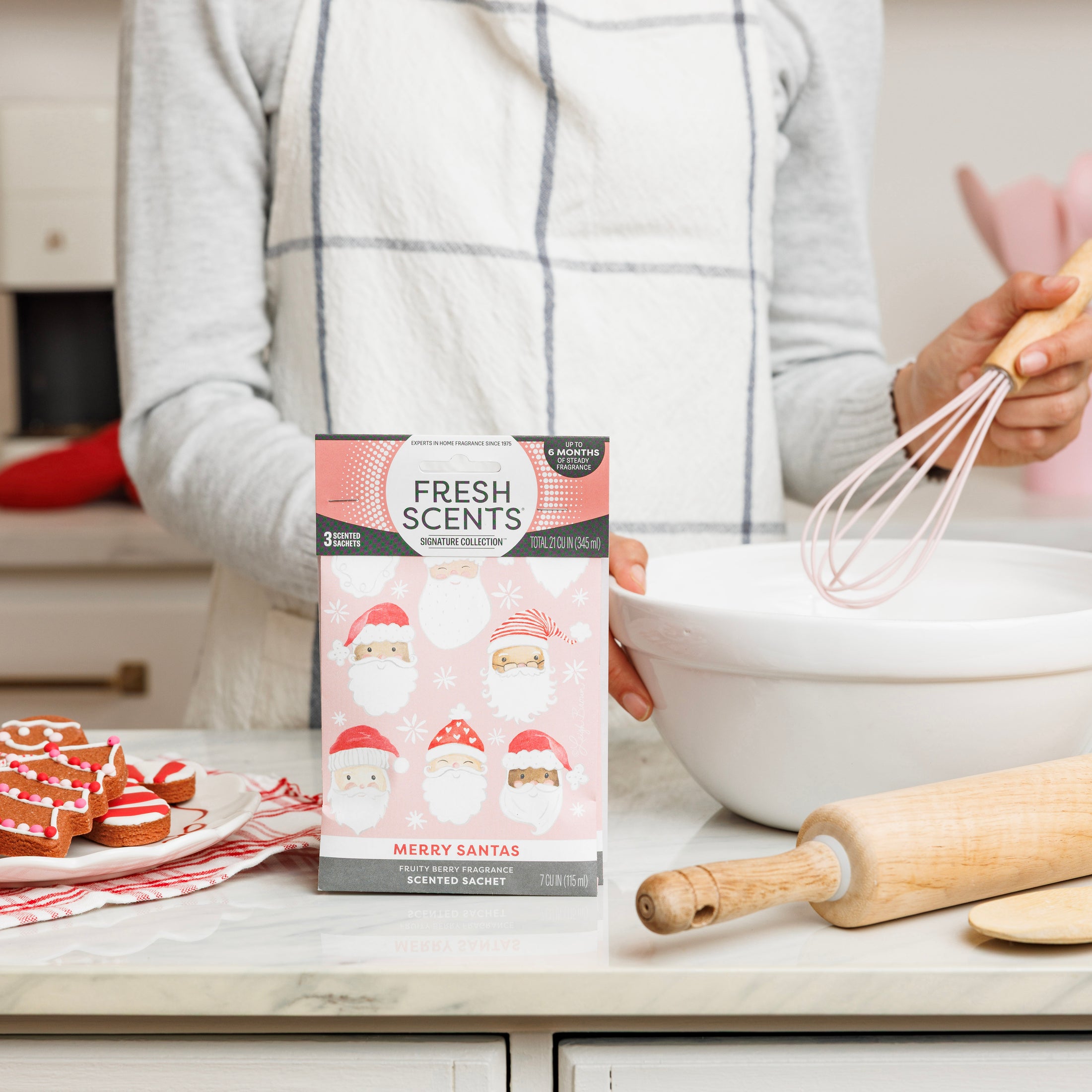 A person in a white apron whisks batter in the kitchen, with decorated Santa cookies, a rolling pin, and a Fresh Scents Merry Santas - Sachet 3pk box for flameless fragrance on the counter.
