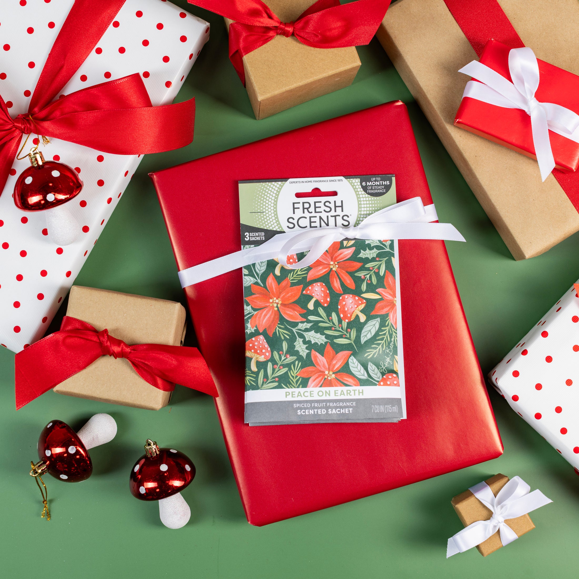 Gift-wrapped presents with red and white ribbons surround a sachet labeled Fresh Scents Peace on Earth - Sachet 3pk, featuring a floral design, all arranged on a green background with decorative red mushroom ornaments. This beautifully displayed set from Fresh Scents serves as a flameless fragrance and long-lasting freshener, making it the perfect addition to your festive décor.