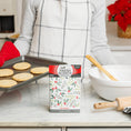Load image into Gallery viewer, A person in a gray shirt and plaid apron holds a cookie tray in the kitchen. In front, the Fresh Scents Be Merry - Sachet 3pk provides long-lasting fragrance near a mixing bowl and rolling pin.

