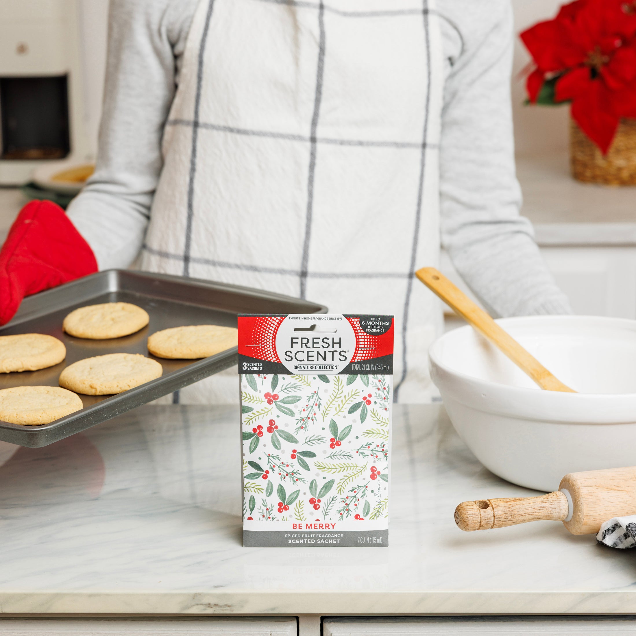 A person in a gray shirt and plaid apron holds a cookie tray in the kitchen. In front, the Fresh Scents Be Merry - Sachet 3pk provides long-lasting fragrance near a mixing bowl and rolling pin.