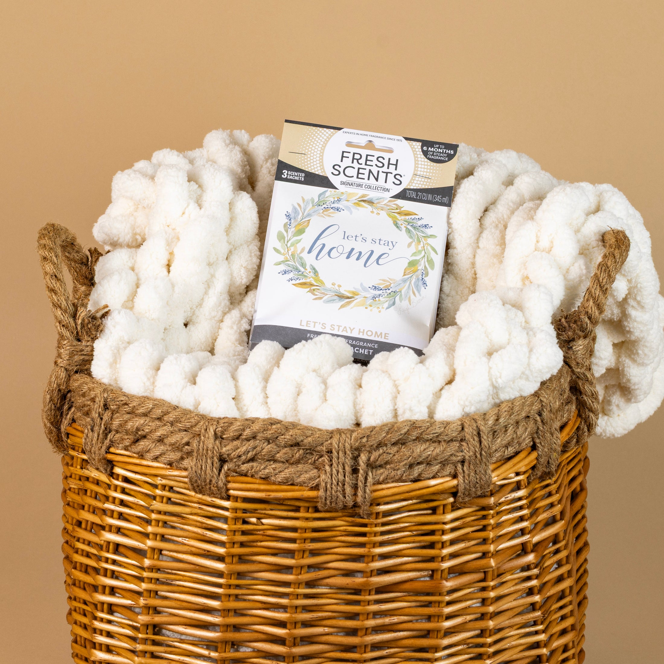 A woven basket holds a chunky white knit blanket and a Fresh Scents Lets Stay Home - Sachet 3pk, a long-lasting flameless air freshener, set against a beige background.