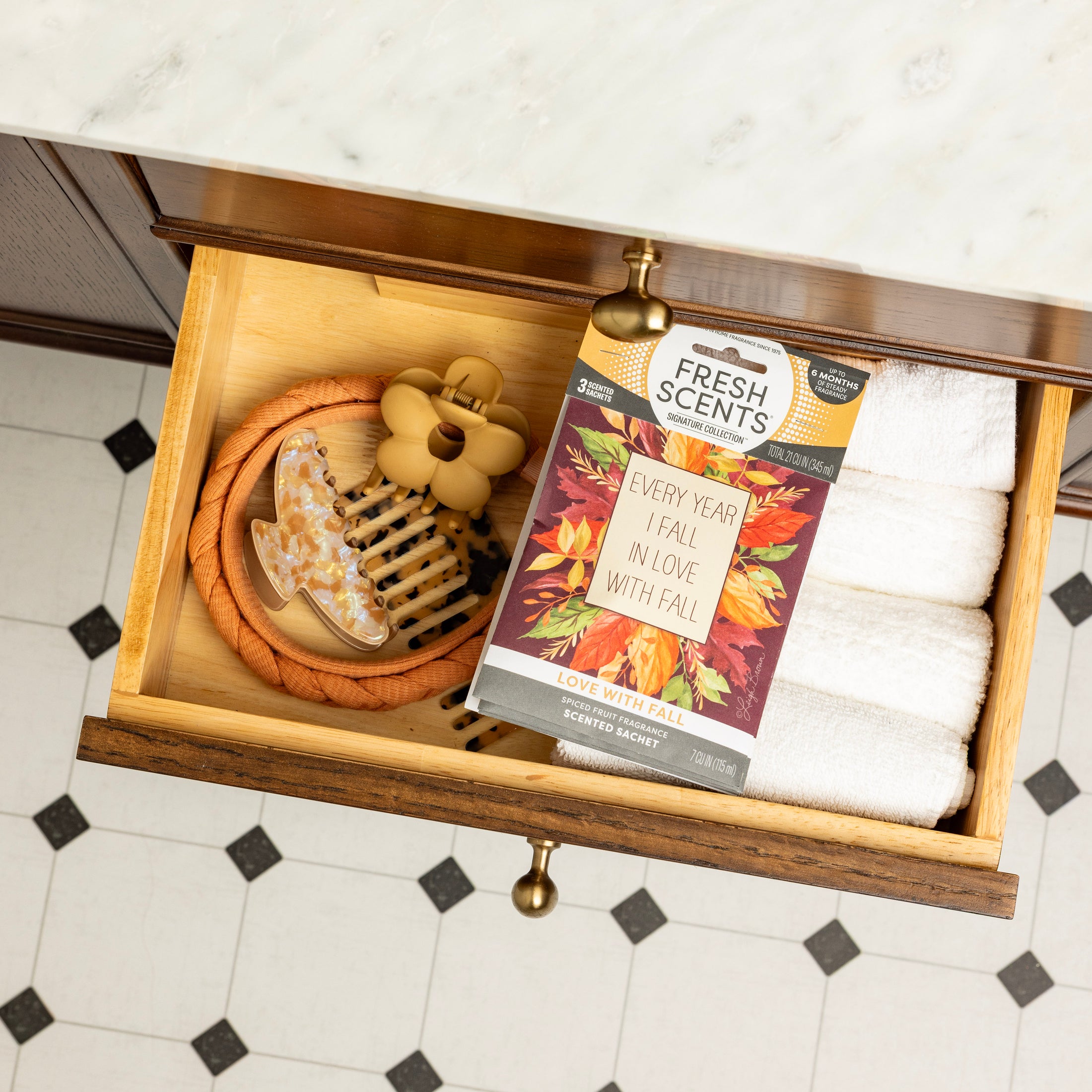 Open bathroom drawer with neatly folded white towels, a braided rope, and Fresh Scents' Love with Fall - Sachet 3pk—a decorative box of autumn leaf-scented sachets—plus a wooden massager, honey dipper, marble countertop, and tiled floor.